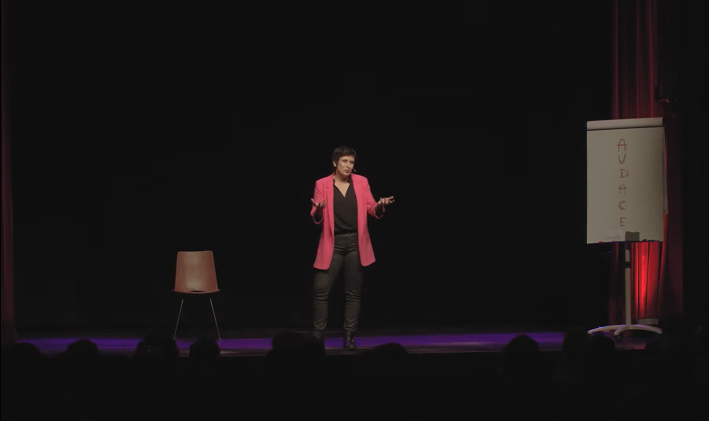 Emilie Somers sur scène, en veste rose, à côté d'un tableau où est écrit Audace.